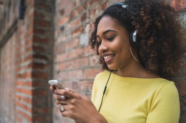 Genç Afro-Amerikan kadın portresi cep telefonu kullanıyor ve sokakta kulaklıkla müzik dinliyor. Açık havada..