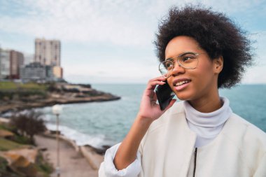 Açık havada, arka planda deniz varken telefonda konuşan genç ve güzel bir Latin kadının portresi. İletişim konsepti.