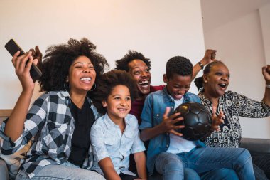 Mutlu Afro-Amerikan çoklu nesil ailesi evdeki oturma odasında televizyonda futbol maçı izliyor. Aile ve spor konsepti.