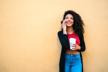 Sarı arka planda bir fincan kahve tutarken telefonla konuşan genç afro kadın portresi. İletişim konsepti.