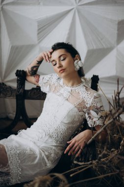 boho tarzında beyaz elbise giyen gelin ahşap bankta oturan ve başında el tutarak   