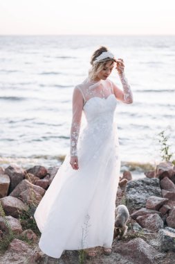 Gün batımında kayalık plajda duran uzun beyaz elbiseli gelinin tam uzunlukta çekilmiş fotoğrafı 
