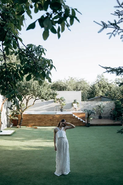 arka planda yüzme havuzu ile yeşil bahçede duran beyaz elbiseli çekici kadın 