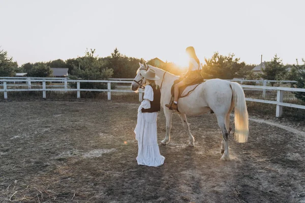 woman holding bridle with girl sitting on white horse at the sunset