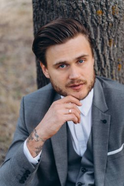 gövde ağacının yakınında yerde oturan gri takım elbiseli çene yakın el ile yakışıklı adam