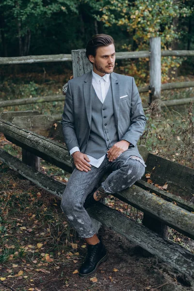 handsome man in gray suit sitting on old wooden fence against forest