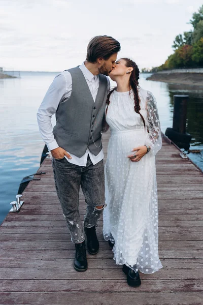 美丽的孕妇在白色礼服与英俊的男人亲吻在湖的木码头 — 图库照片