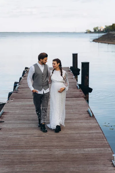 美丽的孕妇在白色礼服与英俊的男人站在一个木码头对湖 — 图库照片