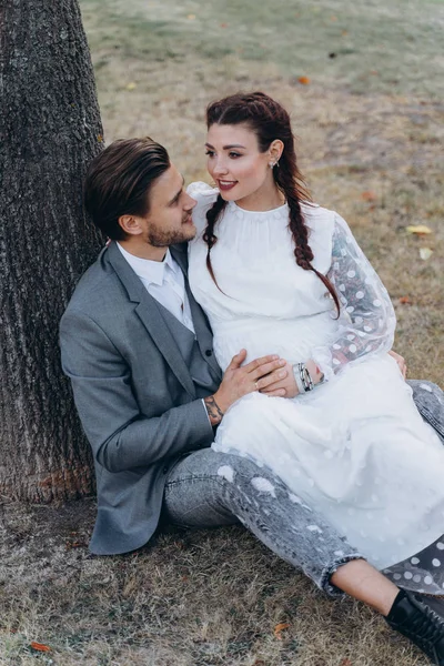 美丽的孕妇在白色礼服与英俊的男人坐在地上附近的树干 — 图库照片