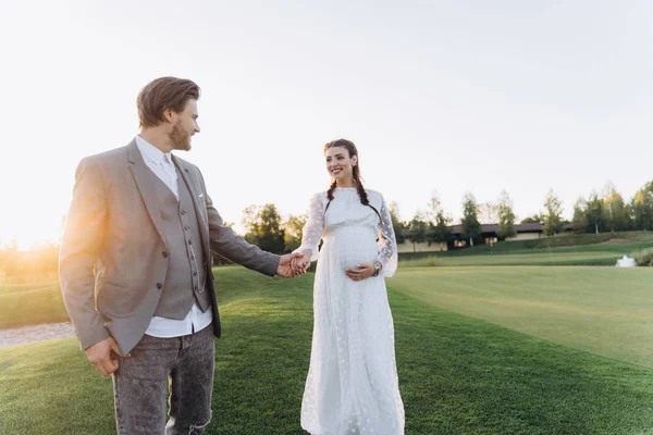 美丽的孕妇在白色礼服与英俊的男人手牵手 走在绿色的草坪上 对夕阳 — 图库照片
