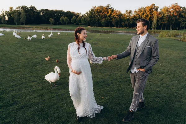 美丽的孕妇在白色礼服与英俊的男人走 并手牵手在绿色的草坪上旁边的白天鹅和鸭子在晚上 — 图库照片
