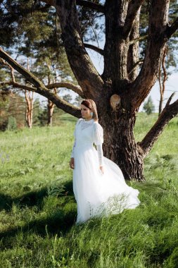 Beyaz elbiseli şık bir kadın çayırdaki ağacın altında duruyor.