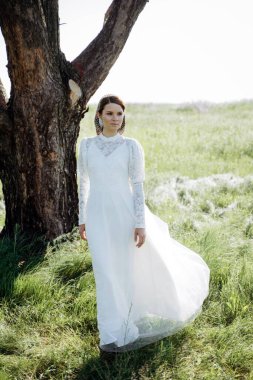 Beyaz elbiseli şık bir kadın çayırdaki ağacın altında duruyor.