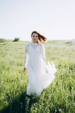 Uzun beyaz elbiseli çekici bir kadın sahada dikiliyor ve kameraya bakıyor. 