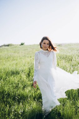 Uzun beyaz elbiseli çekici bir kadın sahada dikiliyor ve kameraya bakıyor. 