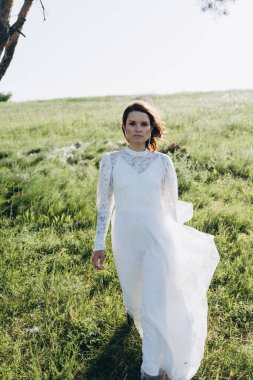 Uzun beyaz elbiseli çekici bir kadın sahada dikiliyor ve kameraya bakıyor. 