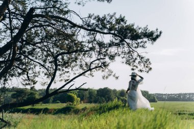 Beyaz elbiseli bir kadının ağaçlı çayırda dikilirkenki görüntüsü. 
