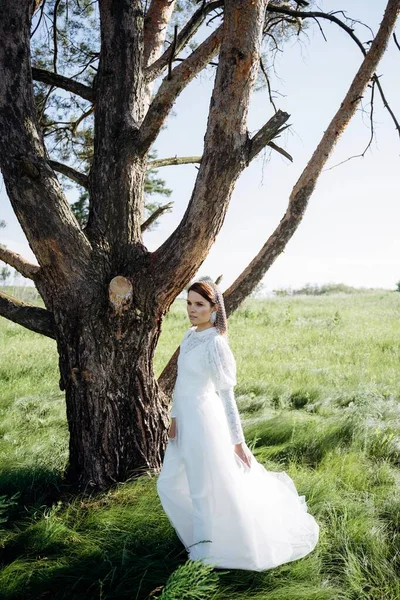 Beyaz elbiseli şık bir kadın çayırdaki ağacın altında duruyor.