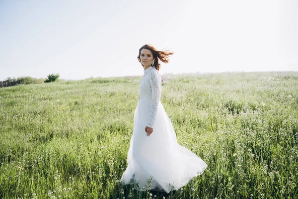 Uzun beyaz elbiseli çekici bir kadın sahada dikiliyor ve kameraya bakıyor. 