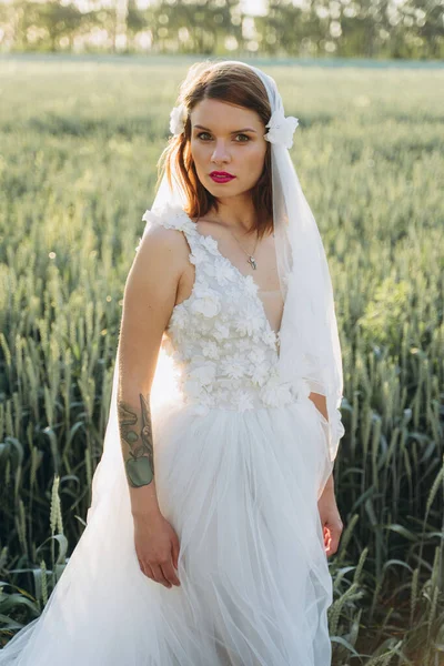 attractive woman wearing veil and long white dress standing in the field, looking at camera