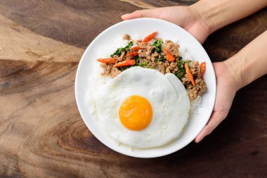 Tayland yemeği, kızarmış kutsal fesleğen kıyma ve sahanda yumurta (Pad Kaprao Moo Kai Dao) ahşap arka planda elle tutulan tabakta