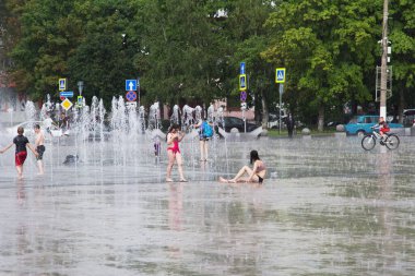 Sıcak bir yaz günü şehir meydanında ıslak giysiler içindeki çocuklar yağmurda asfaltta su birikintilerinde otururlar.