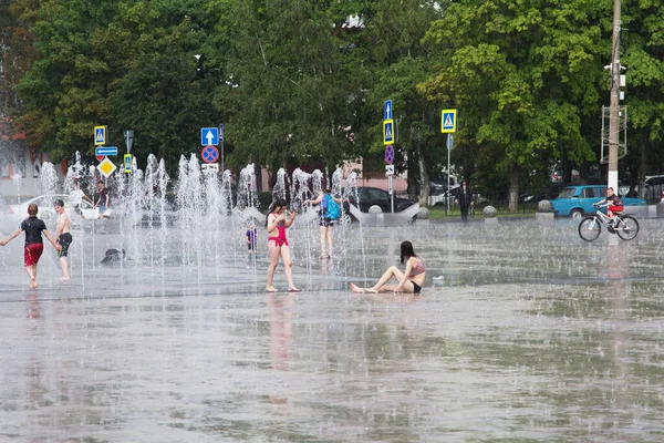 Sıcak bir yaz günü şehir meydanında ıslak giysiler içindeki çocuklar yağmurda asfaltta su birikintilerinde otururlar.