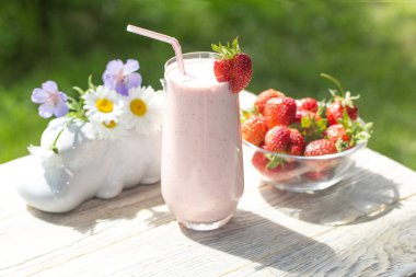 Çilekli milkshake, açık ahşap bir arka planda, arka planda bir vazo kır çiçeğiyle birlikte uzun bir bardakta duruyor.