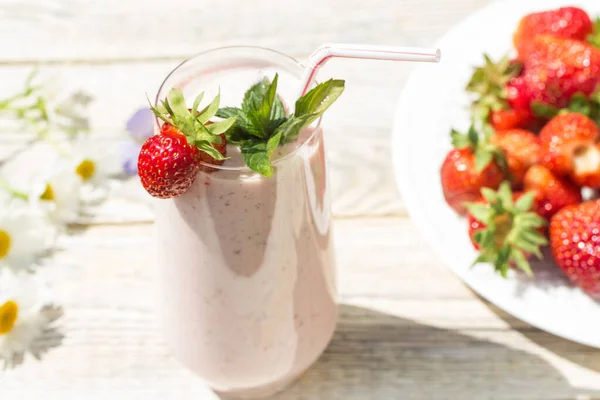 在一个阳光明媚的日子里 在一个轻盈的木制背景上 有一个玻璃杯 上面放着草莓奶昔 一个白色的盘子 上面放着草莓和分散的雏菊 — 图库照片