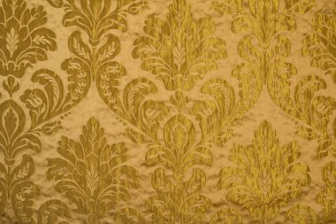 Kumaş deseninde tekrar eden çiçek deseni ve geometrik elementler var. Sarı kumaş rengi. Metalik ipliklerle işlenmiş nakış