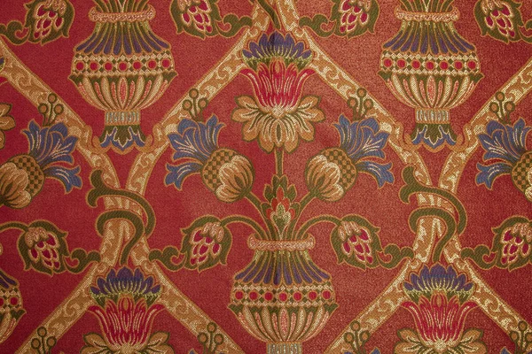 Kumaş deseninde tekrar eden çiçek deseni ve geometrik elementler var. Kırmızı kumaş rengi. Metalik ipliklerle işlenmiş nakış