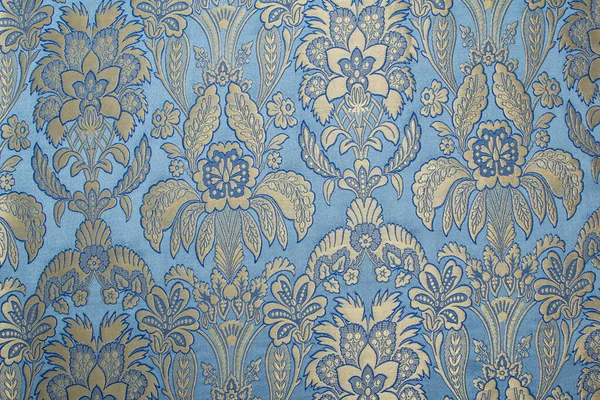 Kumaş deseninde tekrar eden çiçek deseni ve geometrik elementler var. Mavi kumaş rengi. Metalik ipliklerle işlenmiş nakış