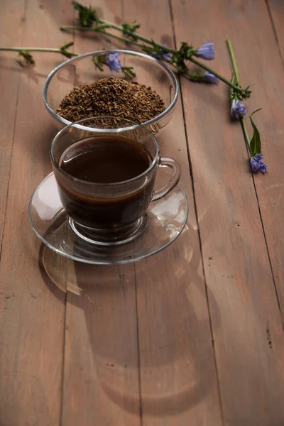 The morning sun shines on an invigorating chicory drink in a glass Cup and a chicory flower on a brown wooden background . Vertical orientation