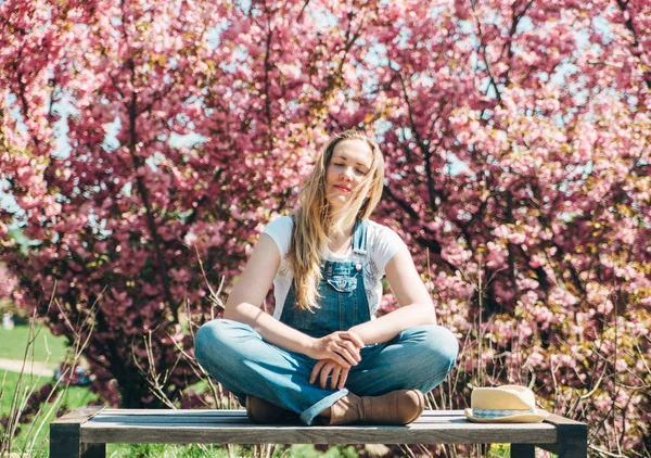 Jonge blonde hipster vrouwen is genieten van tijd buitenshuis. Gelukkig mooi gezicht. Natuurlijke uitstraling. Groene natuur voorjaar achtergrond met roze sakura bloemen. — Stockfoto