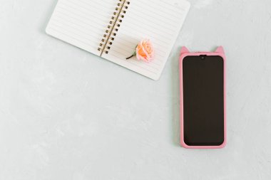 Düz, kadınsı ofis masası. Gri arka planda not defteri, telefon ve çiçek modeliyle.