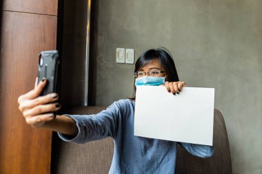Tıbbi maske takan Asyalı bir kız akıllı telefon konferans kamerası kullanıyor ve COVID-19 sırasında elinde boş bir kağıt tutuyor. Sosyal uzaklık ve sağlık konsepti