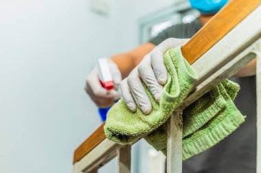 Koruyucu eldivenli genç Asyalı adam evde temizlik yaparken merdiven korkuluklarını dezenfekte ediyor, yakın çekim elleri görüyor ve sprey şişe kullanıyor. Sağlık hizmetleri ve ev konsepti