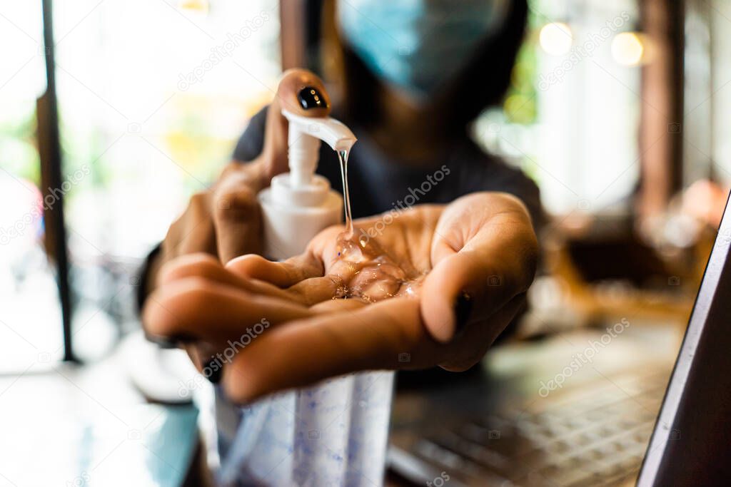 Young woman washing hands with alcohol gel in quarantine for coronavirus wearing protective mask with social distancing and using laptop computer working at home. Work from home and covid-19 concept.