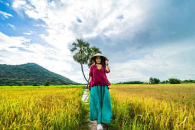 Koyu kırmızı ve yeşil renkli geleneksel kostüm giyen Vietnamlı kız sarı yeşil pirinç tarlasında yürüyen konik şapkalı ve iki palmiye ağacı olan Ba Den Dağı 'nın arka planında mavi gökyüzü. Seyahat kavramı.