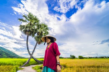 Koyu kırmızı ve yeşil renkli geleneksel kostüm giyen Vietnamlı kız sarı yeşil pirinç tarlasında yürüyen konik şapkalı ve iki palmiye ağacı olan Ba Den Dağı 'nın arka planında mavi gökyüzü. Seyahat kavramı.