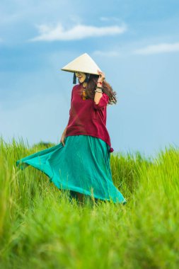 Koyu kırmızı ve yeşil renkli geleneksel kostüm giyen Vietnamlı kız. Sarı ve yeşil pirinç tarlasında yürüyen konik şapkalı. Seyahat kavramı.