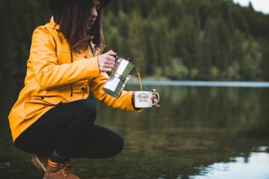  Sarı ceket giyen güzel bir kadın gayzer kahve makinesinden açık hava bardağına kahve döküyor. Doğada sabah içkisi, kampın yakınında kahve hazırlayan bir kız. Macera konsepti başlıyor.