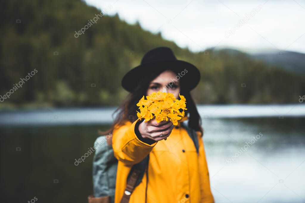 Close up view of female backpacker with hat and yellow jacket handing wild flowers with lake and mountain forest in the background. Flower selected focus. Hikind and outdoor concept. Travel wanderlust