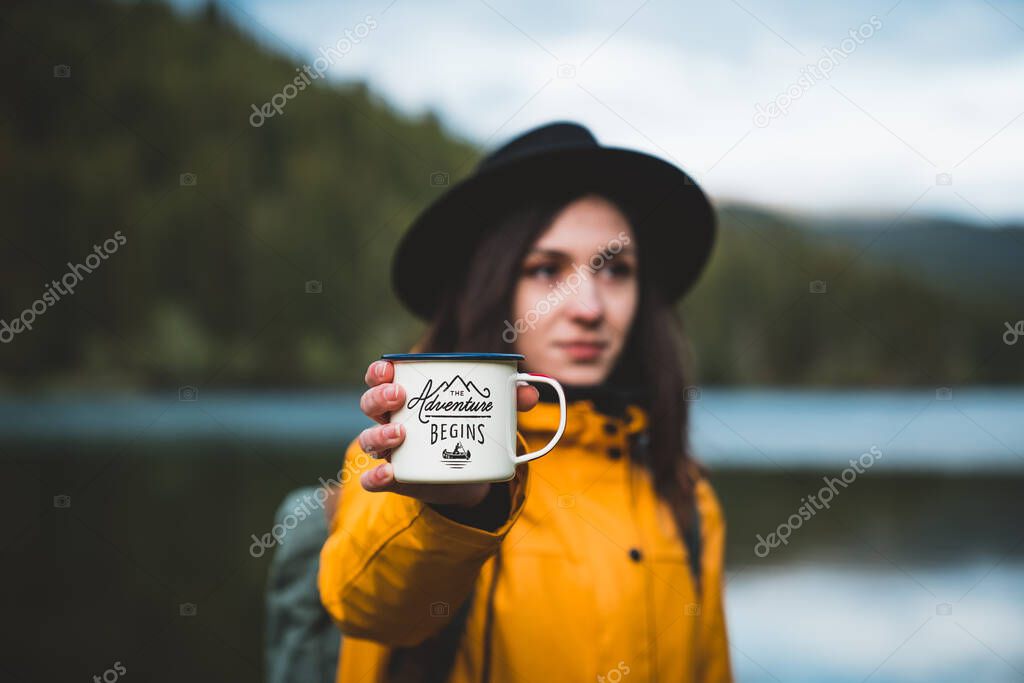 Hipster woman wearing hat and yellow jacket holding outdoor mug or cup. Morning hot coffee in nature. The adventure begins outdoor concept. Travel and wanderlust