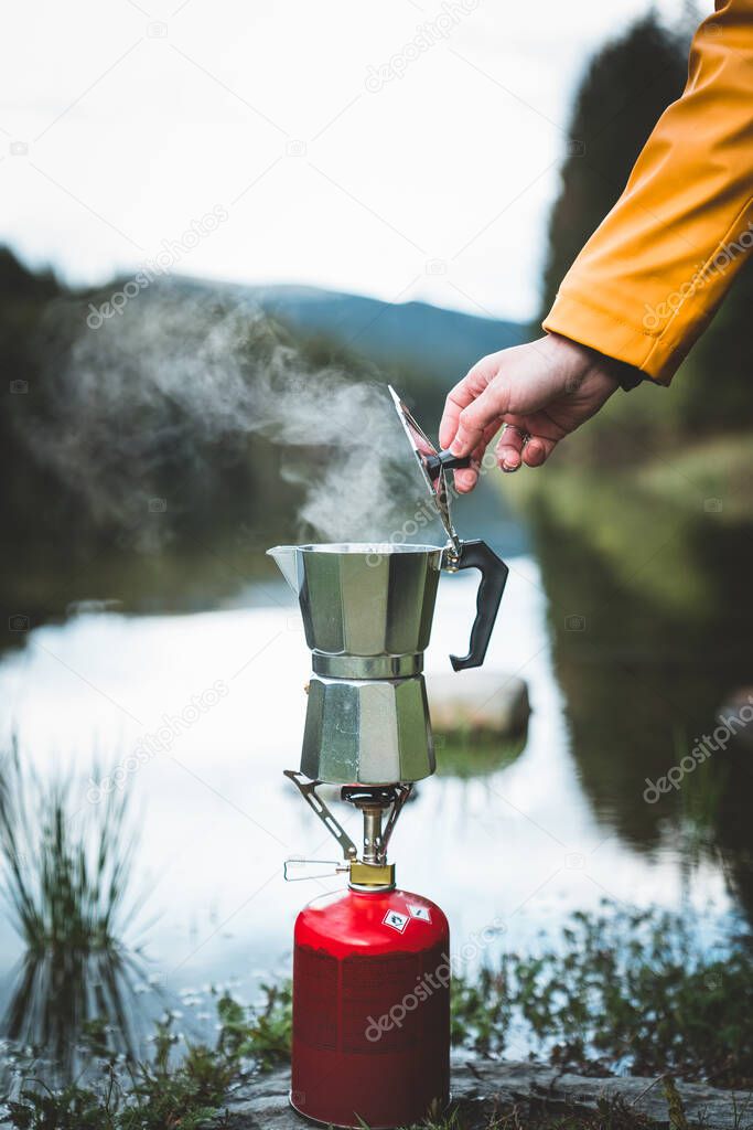 Human hand brewing geyser coffee maker while preparing boiling tea or coffee on camping gas stove by the lake. Camping cooking in nature outdoor. Tourism recreation outside and campsite lifestyle.
