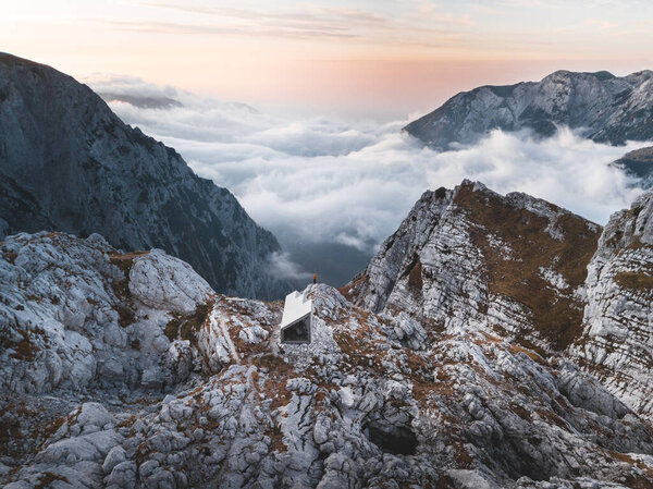 Beautiful aerial photo of misty valley surrounded by rocky mountains. Aerial view of amazing slovenian mountain cabin Bivak Pod Skuto at sunrise. Amazing view above the sea of clouds. Kamnik-Savinja