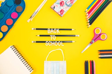 El sanatları malzemeleriyle dolu renkli bir arka plan. Okul mesajları içeren sarı arkaplandaki okul aksesuarlarının görüntüsü. Not defteri, tıbbi maske, makas, kalem ve masanın üzerindeki boya.