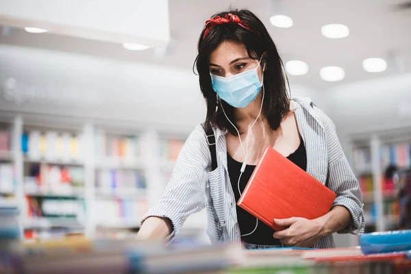 Moda kıyafetleri içinde genç bir kız öğrenci sınava hazırlanıyor ve kütüphanede kitap arıyor. Coronavirus salgını sırasında üniversitede okuyan yüzü tıbbi maskeli sevimli hippi kadın.