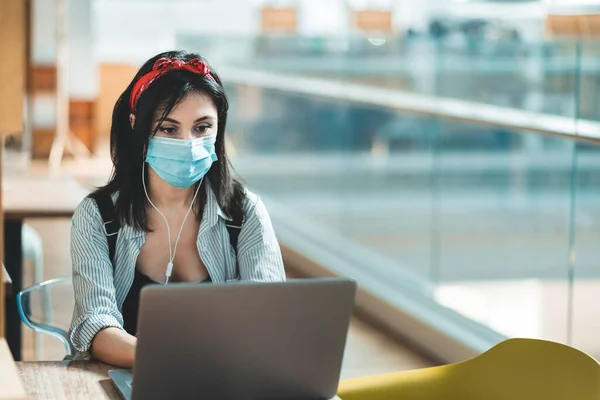 Laptop kullanan, koruyucu yüz maskesi takan ve üniversite kütüphanesinde okuyan genç bir kadın. Kahve dükkanında çalışan bir kadın.
