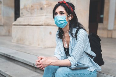 Koruyucu yüz maskesi takan düşünceli genç bir kadın üniversite merdivenlerinde oturuyor ve elleri kelepçeli gelecek için endişeleniyor.
